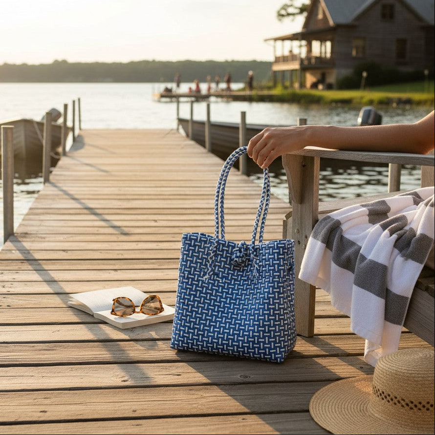 A blue and white woven tote bag held by a person sitting on a wooden dock by the water, with a house and lake visible in the background. A straw hat, sunglasses, and a gray-and-white striped blanket are also on the dock.
