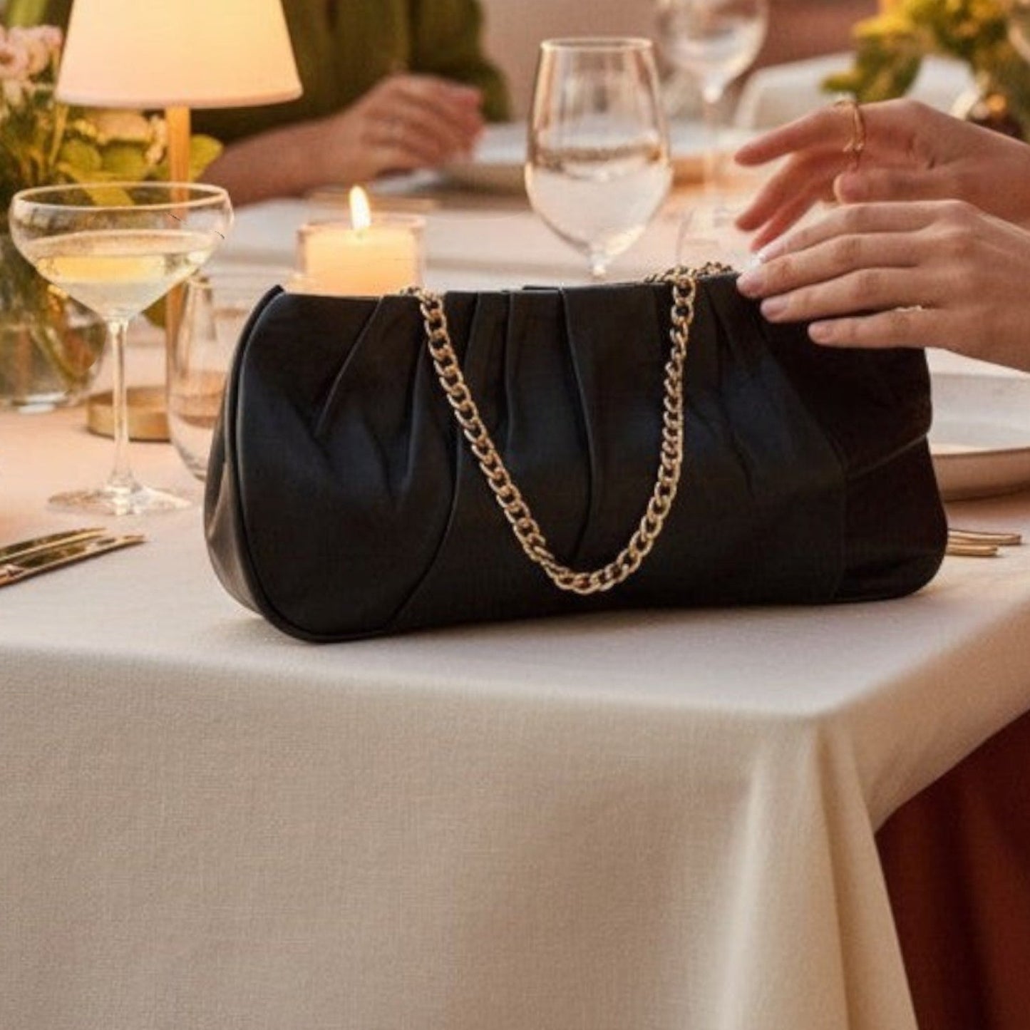 Shop Sarah Stewart Amina black pleated lambskin leather clutch with gold-tone chain strap on a dining table.