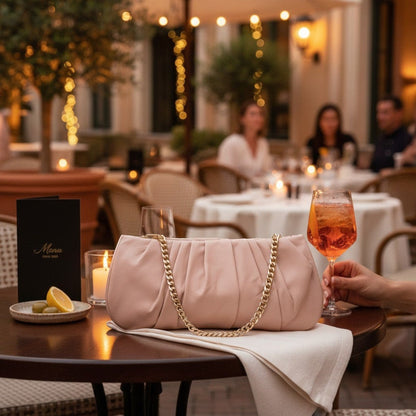 Shop Sarah Stewart Amina pleated blush pink lambskin leather clutch with a gold chain strap on an outdoor cafe table.