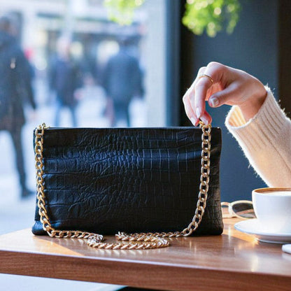 Shop Sarah Stewart Penelope black lambskin leather croc-embossed clutch with gold chain strap on a cafe table