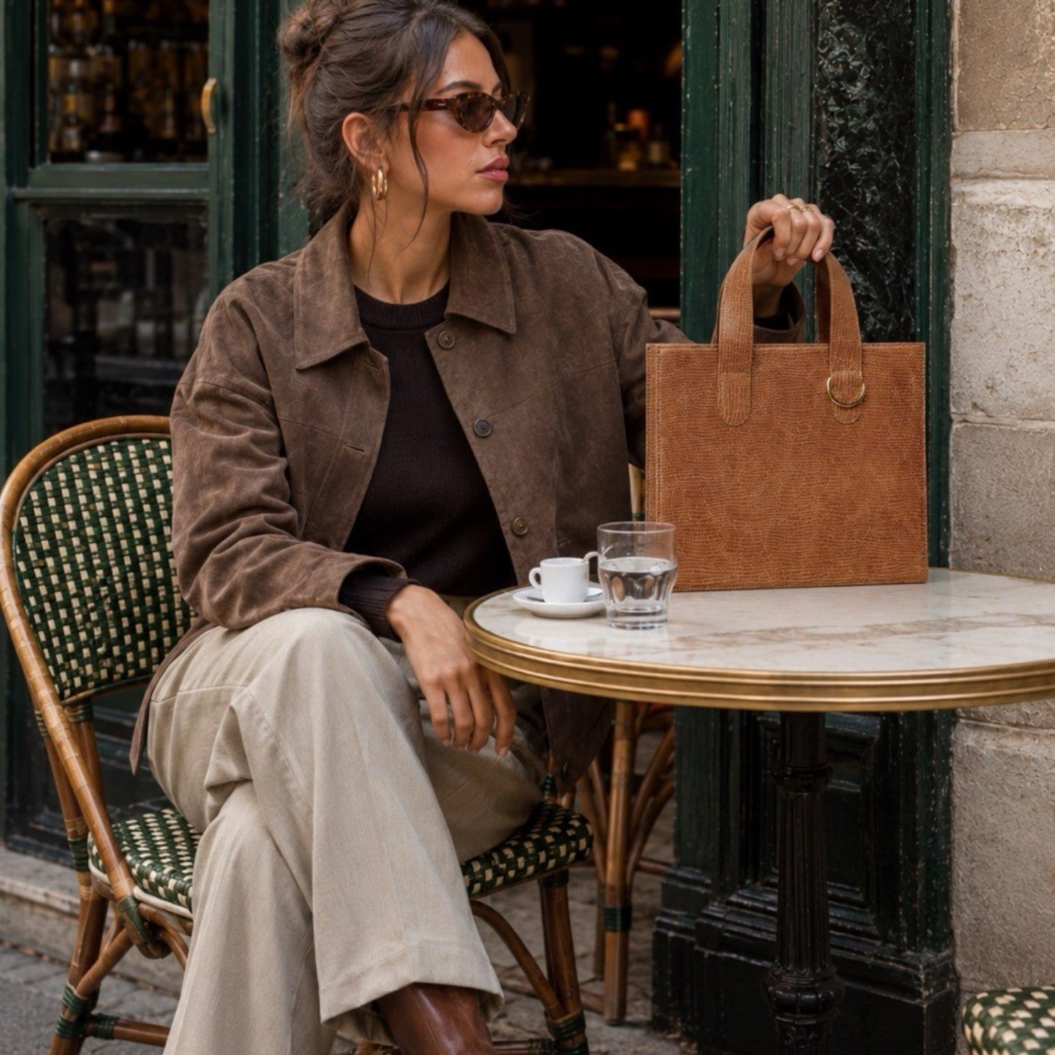 Shop Sarah Stewart Claire Maxi embossed goatskin leather handbag in tan held by a woman at a cafe table.