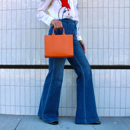 Shop Sarah Stewart orange embossed leather Adelaide handbag held by a woman in a white blouse and flared jeans