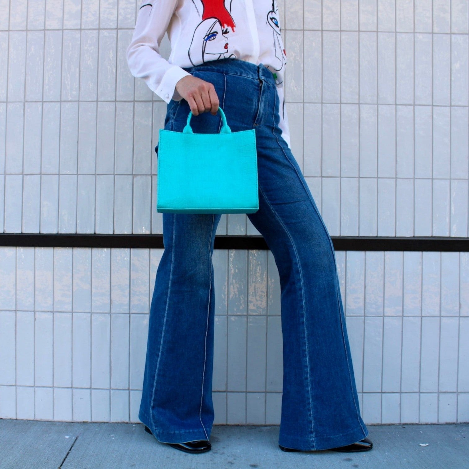 Shop Sarah Stewart Adelaide handbag in bright turquoise embossed goat skin leather held by a model in flared jeans.
