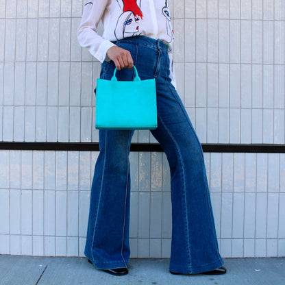 Shop Sarah Stewart Adelaide handbag in bright turquoise embossed goat skin leather held by a model in flared jeans.